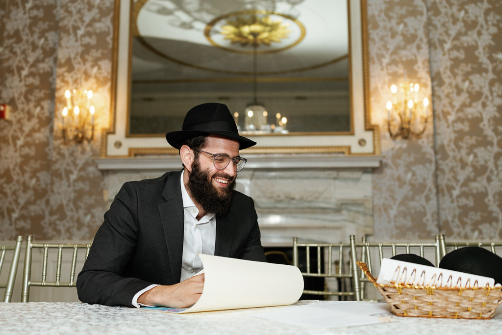 A man wearing a black hat and suit smiles while seated at a wedding event table, reading from documents.